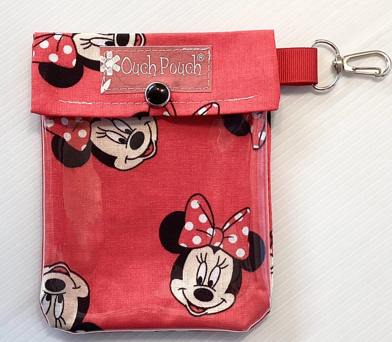 May include: Red fabric pouch with a clear plastic window, featuring a repeating pattern of Minnie Mouse. The pouch has a zipper closure and a small metal loop for attaching a keychain. The text "Ouch Pouch" is printed on the front of the pouch.