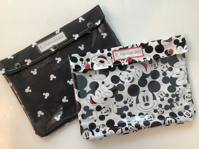 May include: Two clear plastic zippered pouches with black and white Mickey Mouse print. The pouches have black fabric flaps with white Mickey Mouse heads and the name "E. Hernandez" written on each flap.