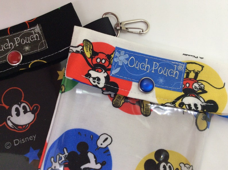 K&ouml;nnte beinhalten: Zwei Mickey Mouse-Themen-Schmerz-Taschen mit durchsichtigen Plastik-Vordertaschen. Die Tasche links ist schwarz mit einem roten Knopf und die Tasche rechts ist wei&szlig; mit einem blauen Knopf und einem roten, gelben und blauen Mickey Mouse-Druck.