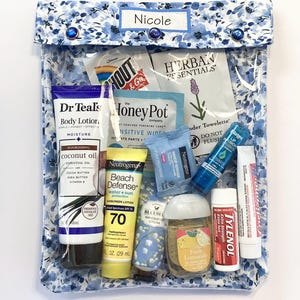 May include: A clear, floral-patterned travel pouch containing various toiletries. Items include lotion, sunscreen, lip balm, hand sanitizer, and makeup remover wipes. The pouch is labeled "Nicole" and secured with blue buttons.