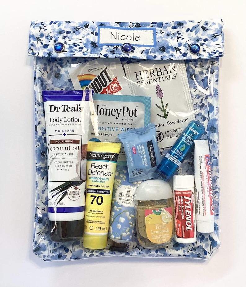 May include: A clear, floral-patterned travel pouch containing various toiletries. Items include lotion, sunscreen, lip balm, hand sanitizer, and makeup remover wipes. The pouch is labeled "Nicole" and secured with blue buttons.