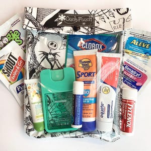 Puede incluir: Una bolsa estampada en blanco y negro con la etiqueta "Ouch Pouch" que contiene varios productos de salud e higiene. Los artículos incluyen protector solar, desinfectante de manos, bálsamo labial, toallitas y analgésicos. Con diseño de Jack Skellington.