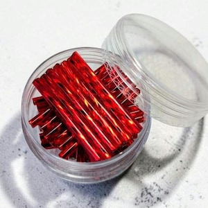 May include: A clear plastic circular container filled with a collection of thin, cylindrical, metallic red sticks. The container has a matching clear plastic lid that is partially open, revealing the vibrant red contents inside the jar.