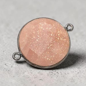 May include: A circular jewelry connector featuring a faceted, peach-colored stone with a shimmering, glitter-like texture. The stone is encased in a dark metallic frame with two small, circular loops extending from opposite sides.