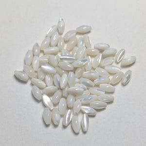 May include: A collection of white, oval-shaped beads with a pearlescent sheen. The beads are scattered across a white surface, creating a delicate and luminous display. These beads are ideal for jewelry making and crafting.