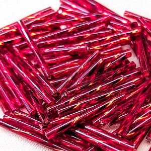 May include: A collection of numerous long, thin, cylindrical glass beads in a vibrant magenta-red color. Each bead features a smooth, reflective surface with a translucent quality that catches the light, creating bright highlights along their length.