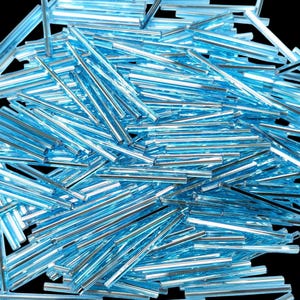 May include: A large collection of light blue, translucent, cylindrical glass bugle beads scattered in a random pile. Each bead features a smooth, elongated, tube-like shape with a reflective surface that catches light, creating a bright, icy appearance.