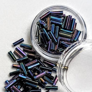 May include: A close-up shot of iridescent, cylindrical beads in a clear container and scattered on a white surface. The beads display a range of colors, including blue, purple, and green, with a metallic sheen. The container is partially open, revealing more beads.
