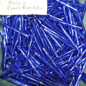 May include: A collection of numerous long, thin, cylindrical blue glass bugle beads with a reflective, glossy finish. A white rectangular label is positioned at the top left corner with the black printed text Made in Czech Republic.
