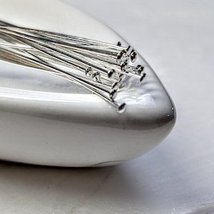 May include: A collection of silver-toned headpins, each featuring a thin, straight metal shaft and a small, flat circular head at one end. These jewelry-making components are arranged in a cluster, showcasing their metallic finish and slender form.