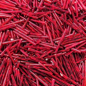 May include: A large collection of long, thin, cylindrical glass bugle beads in a vibrant, deep red color. Each bead features a smooth, reflective surface with a glossy finish, creating a dense, uniform pile of scattered, shiny red tube-shaped pieces.