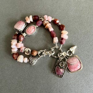 May include: A layered bracelet featuring pink and burgundy gemstone beads. The bracelet includes a variety of shapes and sizes of beads, with silver-toned metal accents and charms. Two charms are visible, one with a heart and the other with a square pink stone.