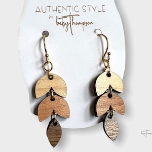 May include: A pair of gold dangle earrings with three wooden leaf-shaped charms. The charms are layered in shades of brown and beige. The earrings are labeled "AUTHENTIC STYLE by betsythompson".