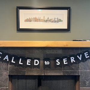 May include: A framed watercolor print of a cityscape hangs above a fireplace. A black banner with white letters that read "CALLED to SERVE" is draped across the fireplace. The wall is painted a muted green.