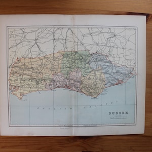 Puede incluir: Un mapa antiguo de Sussex, Inglaterra, con carreteras, pueblos y características geográficas detalladas. El mapa está impreso en papel envejecido con una paleta de colores descoloridos, incluyendo tonos de verde, amarillo y azul. Un bolígrafo negro descansa sobre la superficie de madera.