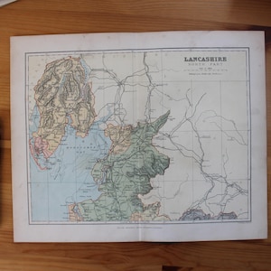 May include: A vintage map of Lancashire, North Part, with detailed geographical features in green, blue, and pink hues. A black pen rests beside the map on a wooden surface. The map is a historical document.