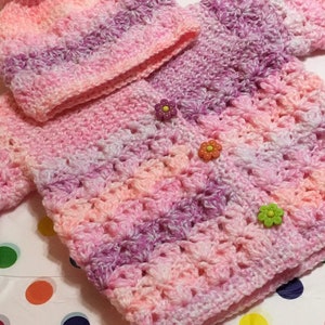 May include: A pink and purple crocheted baby sweater and hat set with flower-shaped buttons.