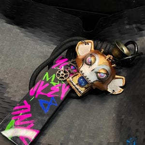 May include: A black and white keychain with a pink, blue, and green graphic that says "Mikey Mouse". The keychain also features a brown, white, and blue robot-like figure with a gear on its chest.
