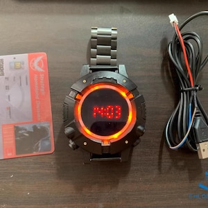 May include: A black wristwatch with a red digital display showing the time 14:03. The watch has a black metal band and a round face with a red ring around the display. The watch is connected to a black USB cable.