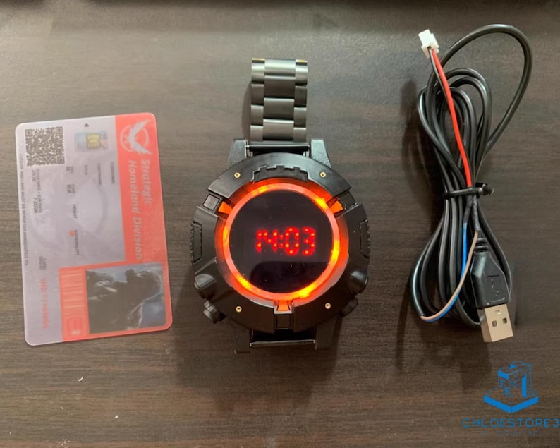 May include: A black wristwatch with a red digital display showing the time 14:03. The watch has a black metal band and a round face with a red ring around the display. The watch is connected to a black USB cable.