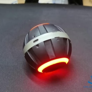 May include: A black and silver spherical Bluetooth speaker with a red light ring around the bottom. The speaker has a textured surface and a small port on the side.