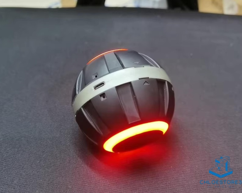 May include: A black and silver spherical Bluetooth speaker with a red light ring around the bottom. The speaker has a textured surface and a small port on the side.