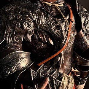 May include: Detailed close-up of a medieval-style suit of armor. The armor is primarily silver and bronze with intricate embossed designs. A leather strap and rope detail the neck area. The armor appears to be a costume or display piece.