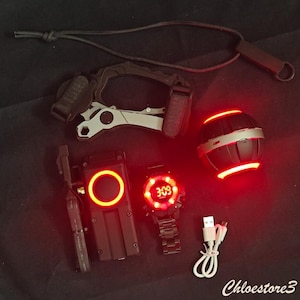 Puede incluir: Un conjunto de equipo t&aacute;ctico negro y plateado, que incluye una multiherramienta, un reloj digital que muestra "3:09", un dispositivo rojo brillante y un objeto esf&eacute;rico con luces rojas. Tambi&eacute;n hay un cable USB blanco.