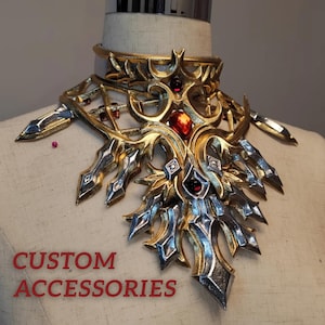 May include: An elaborate gold and silver necklace featuring red gemstones. The necklace has a detailed, layered design with sharp, pointed edges and the text "CUSTOM ACCESSORIES" in red.