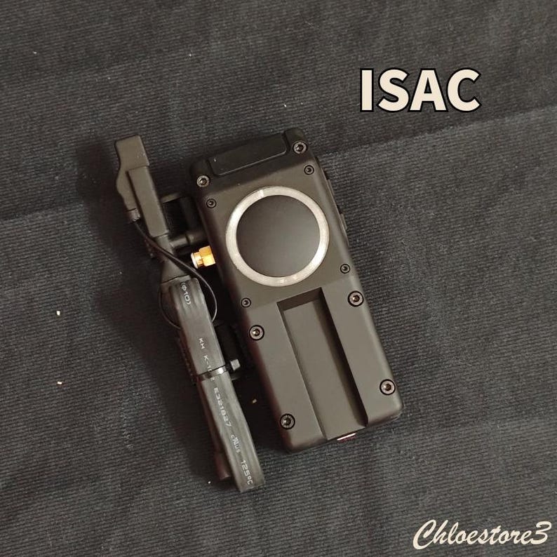 Puede incluir: Dispositivo electr&oacute;nico negro con un dise&ntilde;o circular y la palabra "ISAC" en blanco. El dispositivo tiene forma rectangular con varios tornillos y un componente m&aacute;s peque&ntilde;o adjunto. La imagen est&aacute; sobre una superficie gris oscuro.