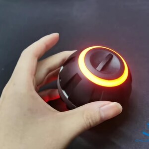 May include: A black and orange round speaker with a glowing orange ring around the top. The speaker is being held in a hand.