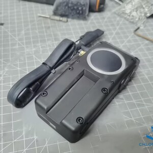 May include: A black electronic device with a circular button and a black cable attached. The device has several screws visible on the side.
