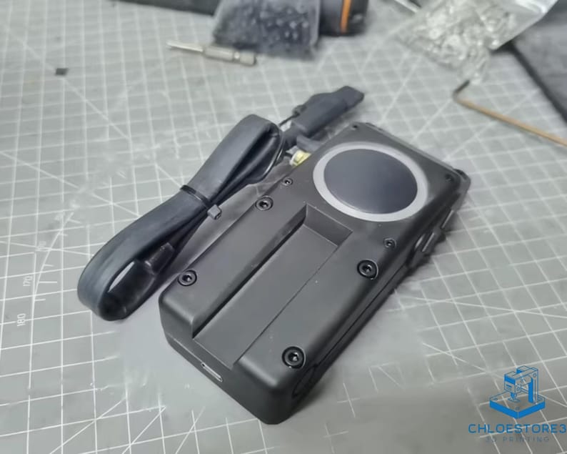 May include: A black electronic device with a circular button and a black cable attached. The device has several screws visible on the side.