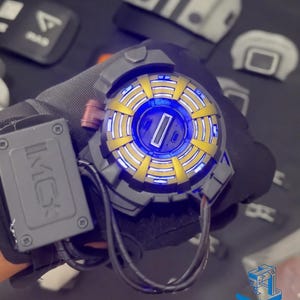May include: A close-up of a black and gray wearable device with a circular, glowing blue and gold center. The device has a black cord attached to it and the text "IKKO" is visible on the side.
