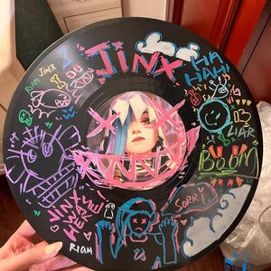 May include: A black vinyl record with colourful drawings and writing on it. The writing includes the words "Jinx", "Rian", "Sorry", "Boom", "Liar", and "Ha Ha Ha".