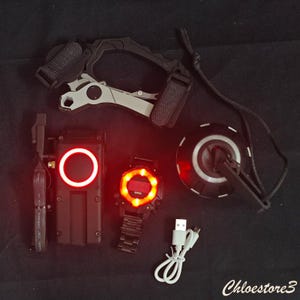Puede incluir: Una colecci&oacute;n de dispositivos tecnol&oacute;gicos negros y grises con acentos iluminados en rojo. Incluye un reloj, un dispositivo de comunicaci&oacute;n, un dispositivo montado en la cabeza y un dispositivo circular con un clip. Tambi&eacute;n hay un cable de carga USB.
