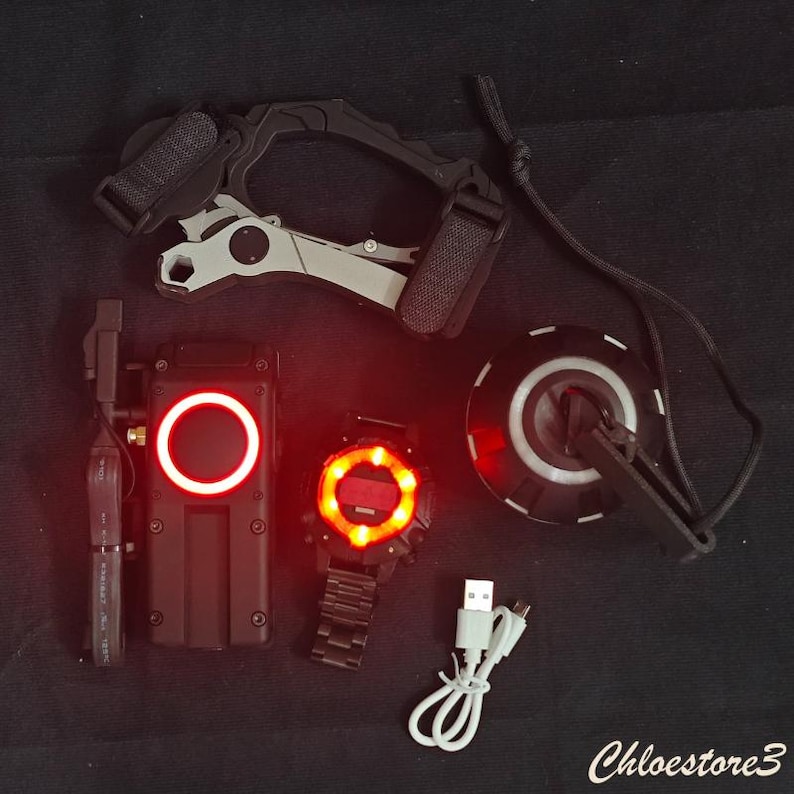 Puede incluir: Una colecci&oacute;n de dispositivos tecnol&oacute;gicos negros y grises con acentos iluminados en rojo. Incluye un reloj, un dispositivo de comunicaci&oacute;n, un dispositivo montado en la cabeza y un dispositivo circular con un clip. Tambi&eacute;n hay un cable de carga USB.