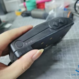 May include: A black handheld device with a textured surface and a circular button. The device has two rectangular buttons on the top and a series of small, round, gold-colored dots on the side.