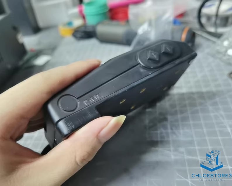 May include: A black handheld device with a textured surface and a circular button. The device has two rectangular buttons on the top and a series of small, round, gold-colored dots on the side.