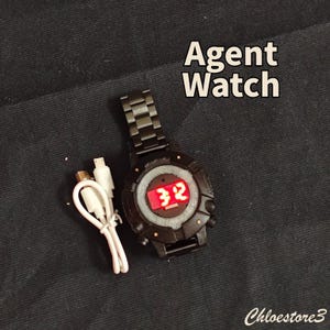 Puede incluir: Reloj Agent negro con una pantalla digital roja que muestra el n&uacute;mero 32. El reloj tiene una correa de metal negro y un cable de carga blanco. El texto "Agent Watch" se muestra en blanco.