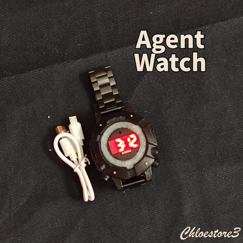 Puede incluir: Reloj Agent negro con una pantalla digital roja que muestra el n&uacute;mero 32. El reloj tiene una correa de metal negro y un cable de carga blanco. El texto "Agent Watch" se muestra en blanco.