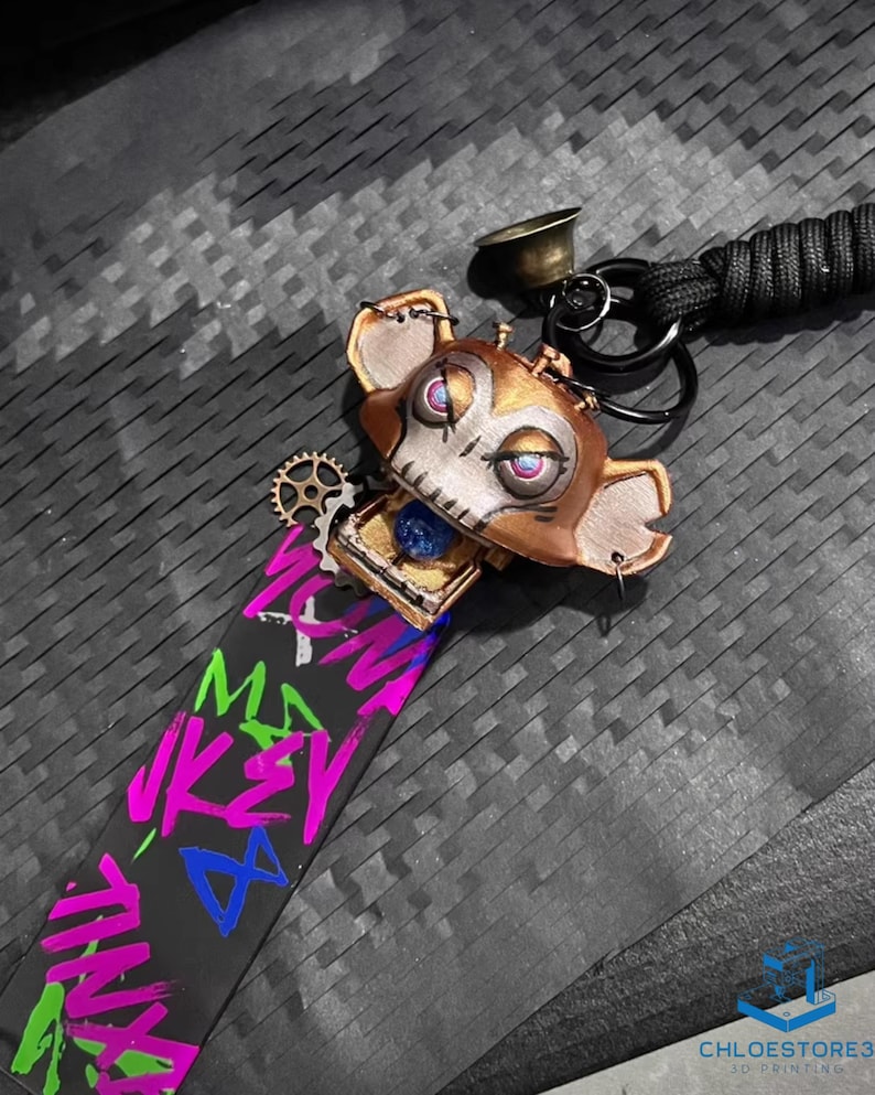 May include: A gold-colored, skull-like keychain with a blue gem in the chest cavity. The keychain is attached to a black strap with colorful graffiti-style text that reads "WOMF MAY JKEY TINY".