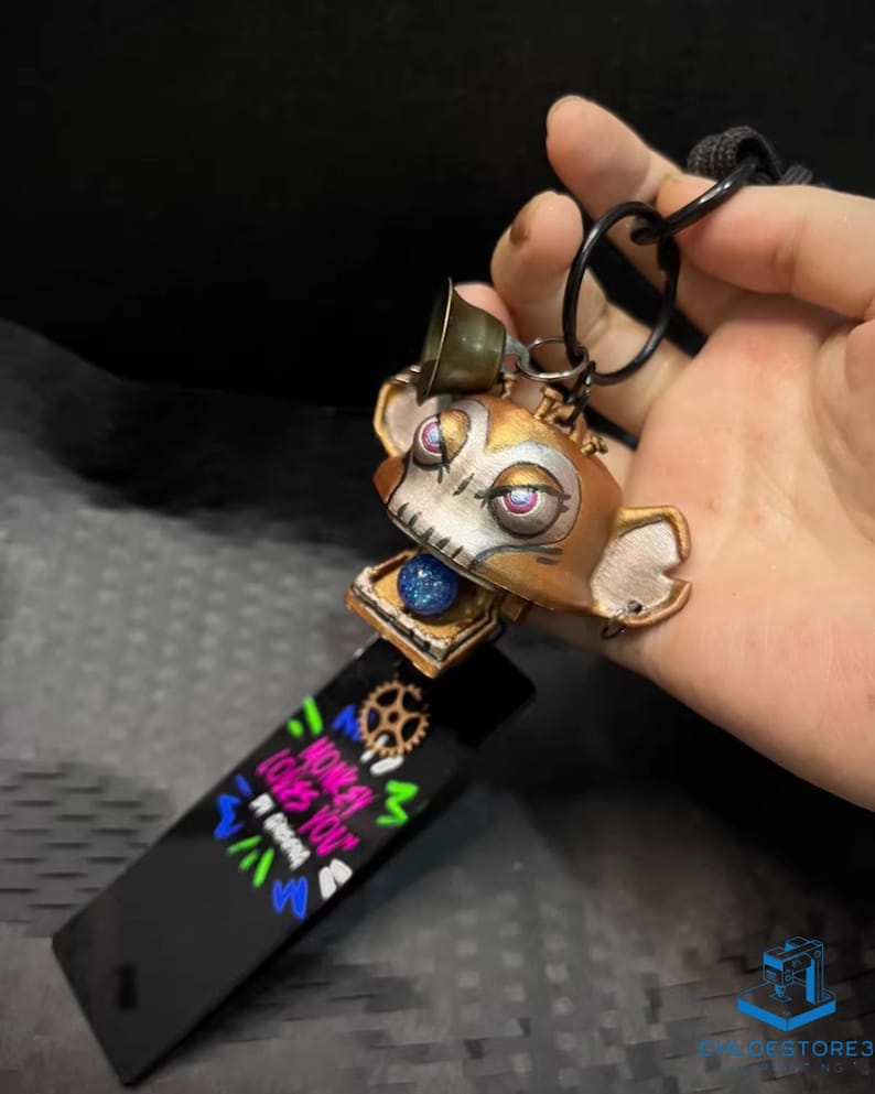May include: A gold and silver robot monkey keychain with a bell on top. The monkey has a skull face with pink and purple eyes. The keychain is attached to a black tag with the words "When you love you" written in colorful letters.