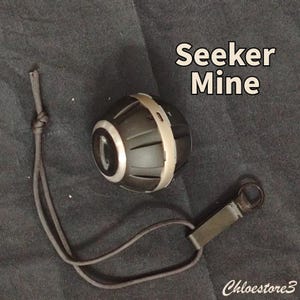 Puede incluir: Un dispositivo Seeker Mine negro y plateado con un cord&oacute;n y un lazo de metal. El dispositivo tiene forma cil&iacute;ndrica con una banda plateada en el medio. El texto "Seeker Mine" se muestra en blanco. El dispositivo est&aacute; sobre una superficie negra.