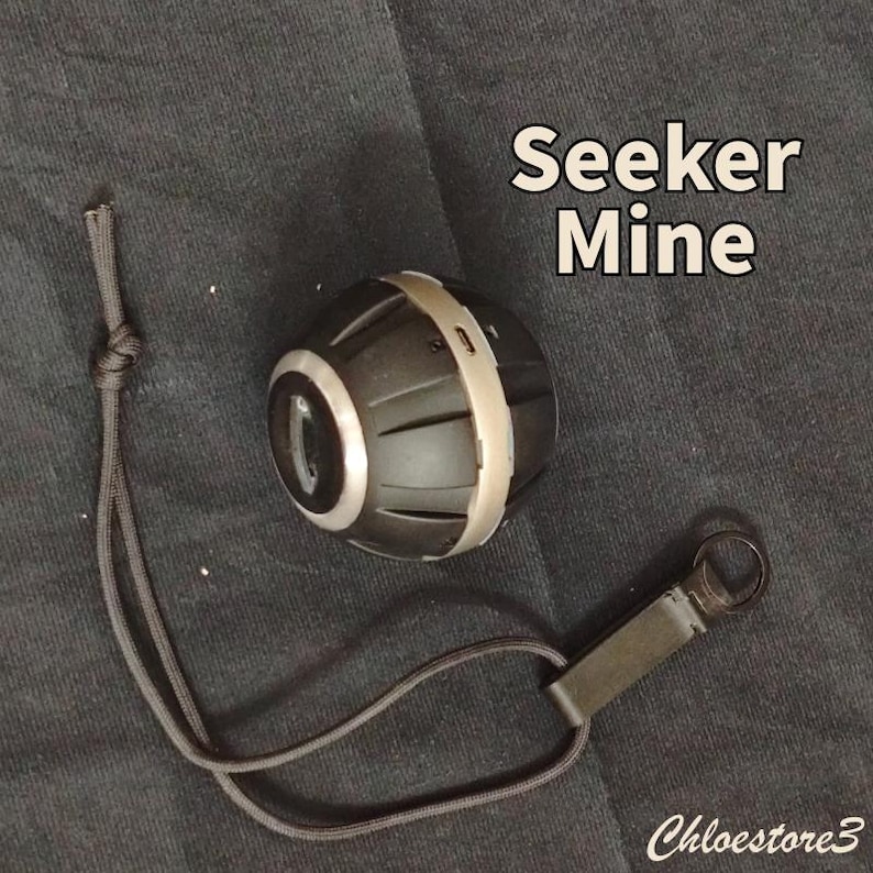 Puede incluir: Un dispositivo Seeker Mine negro y plateado con un cord&oacute;n y un lazo de metal. El dispositivo tiene forma cil&iacute;ndrica con una banda plateada en el medio. El texto "Seeker Mine" se muestra en blanco. El dispositivo est&aacute; sobre una superficie negra.