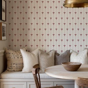 May include: A cozy dining nook with a round wooden table and two wooden chairs. The built-in bench is adorned with various neutral-toned pillows. The wallpaper features a repeating pattern of red floral accents on a cream background. A brass pendant light hangs above.