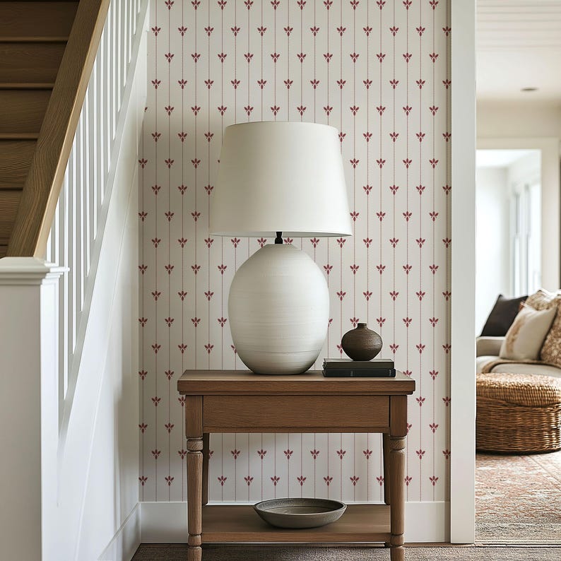 May include: A wooden side table with a white lamp, a small vase, and a decorative bowl. The wall behind the table has a cream-colored wallpaper with a repeating pattern of red floral motifs. A staircase is visible on the left.