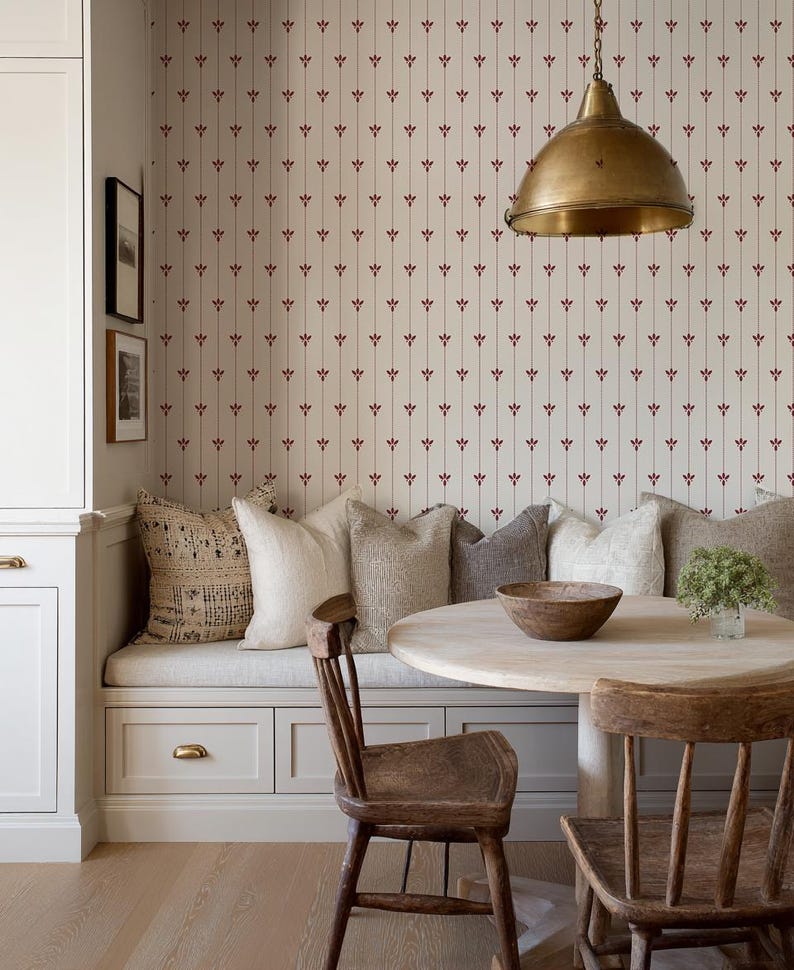 May include: A cozy dining nook with a round wooden table and two wooden chairs. The built-in bench is adorned with various neutral-toned pillows. The wallpaper features a repeating pattern of red floral accents on a cream background. A brass pendant light hangs above.