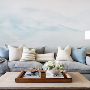 Peut inclure: Un canapé gris orné de coussins blancs et bleus. Une table basse en bois présente un plateau avec des livres, un objet décoratif et un bol blanc de fleurs. Le mur derrière est orné d'une peinture murale à l'aquarelle.