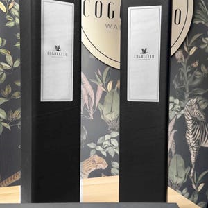 May include: Black rectangular boxes with white labels featuring the "COGOLLETO WALLPAPER" logo. The background showcases a wallpaper design with a jungle theme, including plants and animal illustrations. The boxes are on a light wood surface.